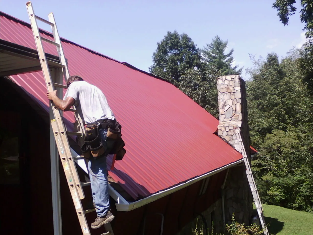 Roofer on a ladder beside a red metal roof and stone chimney — Soffit & Fascia Repair by Roofing Brothers in Rhinelander